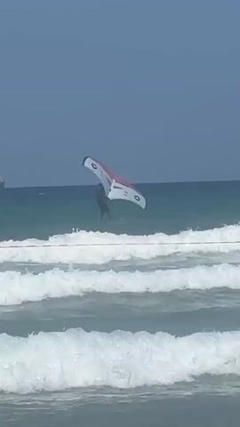 A person is wing surfing on a body of water. The person is in the air above the water, holding onto a wing that is also in the air. The wing is red and white with black accents. The water is blue and green, with white waves. The sky is blue. There are some faint white lines on the water in the foreground. There is a ship in the distance on the left side of the frame.