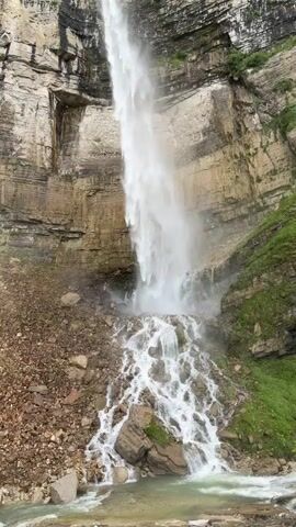 A tall waterfall cascades down a rocky cliff face, disappearing into a mist at the bottom. The cliff is sheer and made of layered rock, with patches of green vegetation clinging to the sides. Below the main waterfall, smaller streams of water flow over a jumble of rocks and boulders. A body of water, likely a river or pool, is visible at the very bottom of the frame, with the water appearing murky and disturbed by the falling water. The overall scene is one of natural power and rugged beauty.