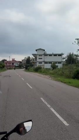 The video is shot from the perspective of someone riding a motorcycle. We see the back of a helmet and a person's shoulder in the foreground. The motorcycle is traveling on a paved road with a white dashed line down the center. On the right side of the road are several buildings, some with balconies, and parked cars. On the left side of the road, there are more buildings and trees. The sky is cloudy. The motorcycle appears to be moving at a moderate speed. The overall environment suggests a rural or suburban setting.