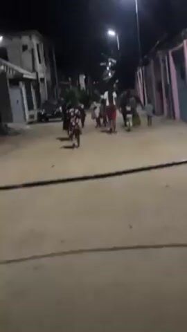 At night, a group of around 10 people, mostly adults, are walking down a dirt road. There appear to be a few children among the group. The street is illuminated by overhead streetlights. There are buildings visible on both sides of the road.  The video then abruptly tilts and the camera appears to be facing the ground and is still.