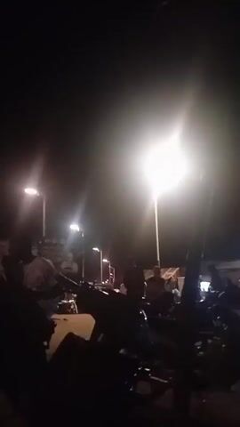 The video is a low-light, shaky recording of an outdoor gathering at night. Several streetlights illuminate the scene, casting a bright glare. Numerous motorcycles are parked in the foreground and midground, with people standing around them. Some individuals appear to be in conversation, while others are seated. The background is indistinct due to the darkness and blurriness. The overall impression is of a casual, informal social event.