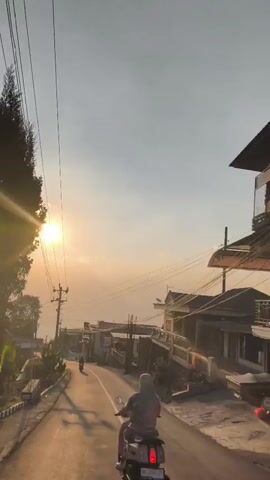 The video shows a person riding a scooter on a paved road during sunset. The sun is low in the sky, casting long shadows and a warm, golden light. There are houses and trees lining the road, and power lines run overhead. The road appears to be in a rural or semi-rural area, possibly on a hill, as indicated by the sloping terrain and distant vegetation. The scooter rider is wearing a headscarf and appears to be alone. The camera is positioned behind the scooter, capturing the scene from the rider's perspective.