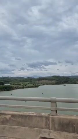 The video shows a view from a bridge looking out over a wide, calm body of water. On the other side of the water, there are rolling green hills covered in trees, with some sparse vegetation and exposed dirt patches on the slopes. In the distance, faint mountain ranges are visible under a cloudy, grey sky. Several small buoys are scattered across the water, and a few small boats or barges can be seen. The foreground of the video is dominated by the concrete structure and metal railing of the bridge, suggesting the viewpoint is from a moving vehicle. The overall atmosphere is somewhat overcast but peaceful.