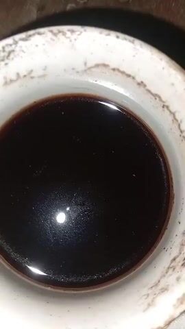 A close-up, top-down view of a dark brown liquid, possibly coffee or tea, in a white ceramic cup with a textured, slightly stained rim. The liquid has a smooth surface with a slight sheen, reflecting a bright light source. The background is out of focus, appearing to be a rough, textured surface.