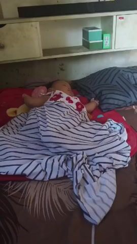 A baby is sleeping on a bed. The baby is wearing a white dress with red flowers and is covered by a blanket with black and white stripes. The bed is covered with a brown patterned sheet and a red blanket is visible underneath the striped blanket. There are pillows on the bed, including a gray and black striped pillow and a pillow with red and blue hearts. In the background, there is a shelf with boxes.