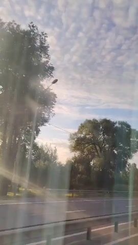 The video shows a roadside view from a moving vehicle. The sky is filled with fluffy white clouds against a blue background. Sunlight streams in from the left, creating lens flare. On the right side of the road, there are trees and a utility pole. A truck passes by in the first few seconds, followed by a car. A billboard and a road sign are visible further down the road.