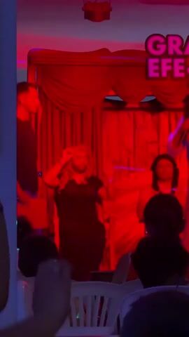 A person wearing a blonde wig and makeup is seated in a chair on a stage. The stage is lit with blue and red lights and has a red curtain in the background. Another person is standing to the left of the seated person. The seated person is moving their arms and head, appearing to be performing.