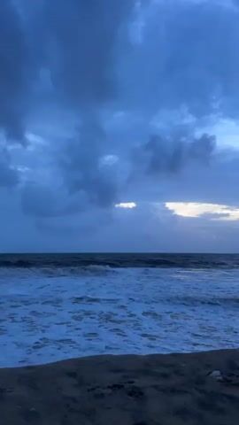 The video shows a view of the ocean with waves crashing on a sandy beach. The sky is cloudy and dark blue, with a sliver of light visible on the horizon. The overall mood is somber and dramatic. There are no people or man-made objects visible in the frame.