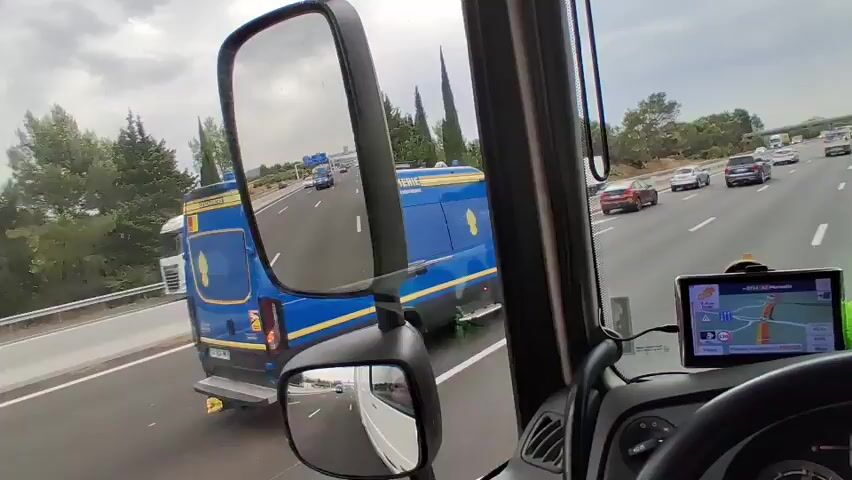 The video is captured from the perspective of a truck driver, looking forward and using the side mirrors. The truck is on a highway with multiple lanes of traffic. In the foreground, the dashboard and a GPS device are visible. The GPS shows a route with a red line indicating the path. In the left side mirror, other vehicles, including trucks and cars, are seen traveling in the same direction. A white pickup truck is in the adjacent lane. In the main forward view, traffic includes cars and vans, one of which is blue and has yellow markings. Trees and vegetation line the side of the highway. The sky is overcast.