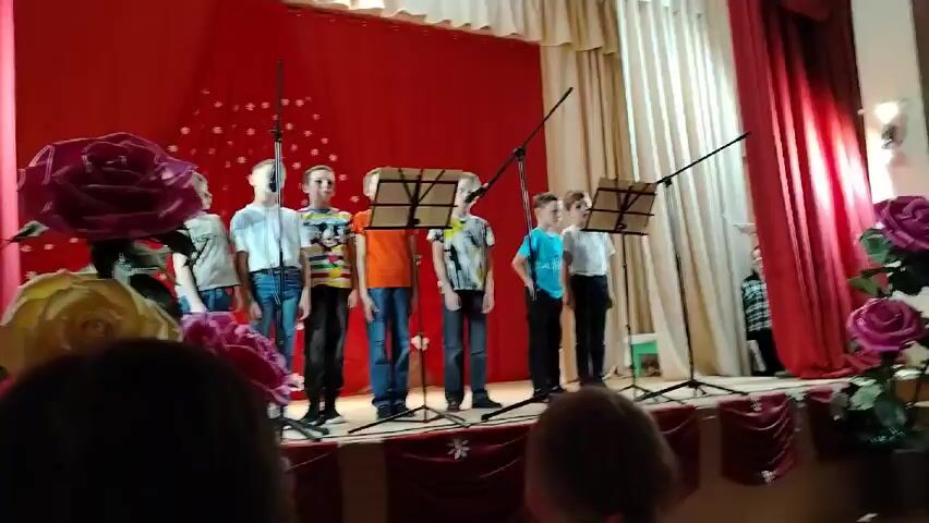 A group of six young boys are singing on a stage decorated with artificial red and yellow roses. They stand in a line behind microphone stands, reading sheet music. The backdrop is a red curtain with white snowflake decorations.  Behind the red curtain is a white curtain.  To the right of the stage, another red curtain hangs down. The audience can be partially seen in the foreground, focusing on the performance.