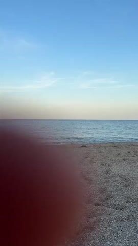 This is a vertical video of a tranquil beach scene at El Parador de las Hortichuelas, Spain. The foreground is a pebbly beach with footprints visible in the sand, suggesting recent activity. The middle ground is the calm, blue sea, with gentle waves lapping at the shore. In the distance, the horizon is a clear line separating the sea from the sky. The sky itself is a gradient of soft blues, transitioning from a paler hue near the horizon to a deeper blue overhead, with a few wispy white clouds scattered across.

The lighting and the soft colors of the sky suggest that the time of day is either early morning or late afternoon, likely during golden hour. The weather appears to be pleasant and clear, ideal for a relaxing beach experience. There are no people or distinct objects visible in the frame, emphasizing the serenity and natural beauty of the location. The overall mood conveyed by the imagery is peaceful and contemplative, inviting the viewer to enjoy the quietude of the coastal landscape. No text is visible in the video.