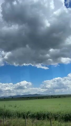 The video shows a green field stretching to a distant line of hills under a sky dominated by fluffy white clouds interspersed with patches of blue. A large mass of dark, grey clouds hangs above the lighter ones, suggesting an approaching storm. The view is framed as if looking out from a window, with faint vertical streaks potentially indicating glass or another transparent surface. The foreground contains tall grass and some scrubby bushes, with a wire fence visible in places. The overall impression is that of a rural landscape, with a contrast between the bright, open field and the looming presence of the darker, heavy clouds.