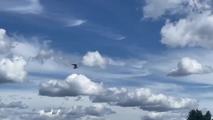 A hang glider with a red and black wing is seen soaring through a blue sky filled with fluffy white clouds. The glider appears to be moving from left to right across the frame. The overall impression is one of a clear, sunny day with favorable flying conditions.