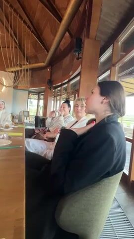The video shows a group of four women sitting around a long wooden table in what appears to be a restaurant or meeting space. The woman in the foreground is wearing a black blazer and black pants. She is speaking and gesturing with her hands. The other women at the table are listening. One is wearing a striped shirt, another is wearing a white blazer, and the fourth, seated to the left, is in a white outfit with a matching hat. There are cups, saucers, and small plates on the table, as well as yellow paper and pencils. The interior of the space features a high ceiling with exposed wooden beams and unique light fixtures. Large windows provide natural light, and there's a view of the outdoors.