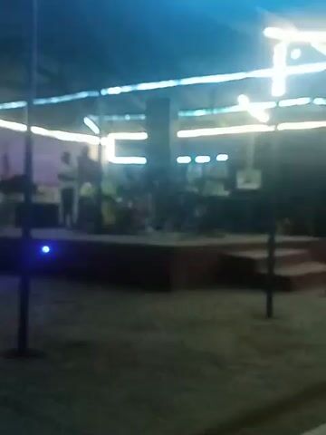 The video shows a band performing on an outdoor stage under a canopy with colored lights. The band consists of at least five members, including vocalists, drummers, and instrumentalists, all dressed in what appears to be white or light-colored shirts. The stage is raised with steps and surrounded by metal railings. A tall speaker stands prominently on the stage. The ground around the stage is sandy or dirt, and there are supporting poles visible. The colored lights above create an ambient glow, changing from green to purple throughout the video. The scene is dark, with lighting primarily provided by the stage lights.