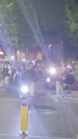 The video shows a nighttime street scene with a crowd of people. The lighting is dim, with streetlights and vehicle headlights illuminating the area. There are trees lining the street, and buildings can be seen in the background. Several people are walking or standing, and some appear to be gathered in groups. There are also some motorbikes present, with their headlights on. Yellow bollards with blue cycling symbols are visible on the road. In the distance, a structure or decoration with rainbow colors is visible above the crowd. Some individuals in the foreground are wearing casual clothing. The overall atmosphere appears to be lively and social.