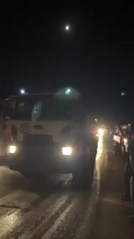 Here is a concise description of the video:

The video is shot at night on a road. A white tanker truck with its headlights on is parked on the side of the road. The tanker has red lights illuminated on the back of the tank. A few people are standing nearby. Other vehicles with headlights on are further down the road in the distance. A bright moon is visible in the sky above. A horn sounds in the video. The road appears wet.