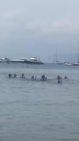 In a medium shot, a group of about a dozen people are in the water, with only their heads and shoulders visible, holding what appear to be blue pool noodles. In the background are several boats, including a larger yacht-type vessel, and a sailboat. The sky is overcast, and the water is calm.