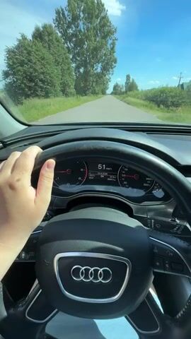 The video captures a first-person perspective from inside a car driving on a road in Dovhe, Ukraine. The dashboard and steering wheel of an Audi are clearly visible, with a hand resting on the wheel. The digital display shows a speed of 51 km/h, suggesting a moderate pace of travel. Outside the car, a bright, sunny day is depicted with a clear blue sky and scattered white clouds. Lush green trees and fields line both sides of the asphalt road, indicating a rural or semi-rural setting. The road ahead appears to be straight and leads towards a horizon dotted with more trees and some distant houses. The overall atmosphere conveyed is one of a pleasant drive through the Ukrainian countryside on a beautiful summer day. The visible text on the dashboard includes "Скорость" (Speed) and "51" which is the current speed, and "28.04.2024" (April 28, 2024) and "09:07" (9:07 AM).