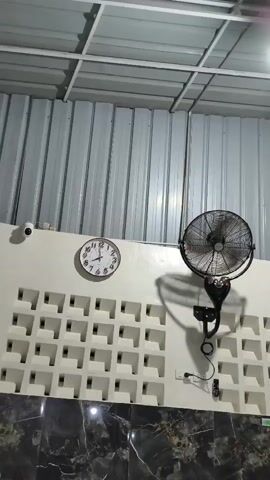A white analog clock with black numbers and hands hangs on a white wall. To its left is a small white security camera. To the clock's right is a black oscillating fan with a protective cage, mounted on a black arm. The fan is plugged into a white electrical outlet. Below the clock and fan is a section of white latticework, with a pattern of square openings. Below the latticework is a section of dark marble with a white veined pattern. Above the clock and fan is a white ceiling with exposed metal beams and ribbed metal siding. The video is a static shot of this scene.