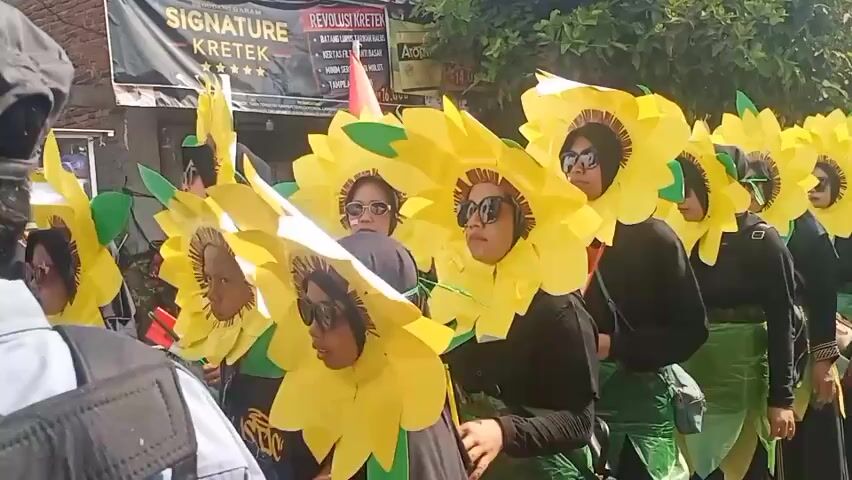 A group of people dressed in sunflower costumes are participating in a parade. They are wearing black shirts with green skirts and large yellow sunflower headpieces. Some are holding Indonesian flags. A man in a security uniform with a cap is walking in front of the group. Another person is filming the parade with a smartphone. The environment appears to be a street with buildings and trees in the background. The overall atmosphere seems festive.