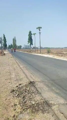 The video shows a paved road with a dry, harvested field on the right side. The sky is clear and blue. On the left side of the road, there are trees and some buildings in the distance. A motorcycle with two riders is visible on the left side of the road. The camera appears to be moving along the road.