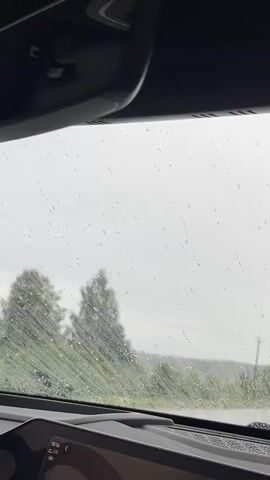 The video captures a view from inside a car on a rainy day. Raindrops streak down the windshield, obscuring the outside view. In the foreground, the car's dashboard is visible, along with a small black robot-shaped air freshener with heart-shaped eyes. Outside, through the rain-streaked glass, a landscape of trees and foliage can be seen. There are power lines and a road visible in the distance, suggesting the car is on a highway or rural road. The sky is overcast and gray, typical of rainy weather. The car appears to be in motion, as evidenced by the streaking raindrops and the slightly blurred distant landscape.