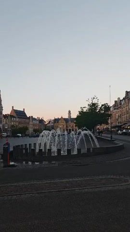 The video captures a serene European town square, likely in Ypres, Belgium, during either early morning or late afternoon, given the clear, pale sky. It opens with a wide view showing a modern fountain with multiple water jets in the foreground. Beyond the fountain, the square is flanked by traditional buildings, culminating in a magnificent, large stone structure with a prominent spired tower on the left side of the frame.

The camera then pans left, focusing on this grand building. It exhibits intricate Gothic architecture with a long facade featuring numerous arched windows and decorative stone carvings. A dark, possibly slate, roof stretches along its length, with several dormer windows and colorful banners hanging from the upper stories.

The pan continues, zooming slightly to highlight the building's towering belfry. This tall, ornate structure features multiple levels, spires, and a large clock face visible near its peak. Small statues or figures are detailed within niches on the tower's facade. The stone appears to be light grey or beige. A red circular traffic sign with a white horizontal bar, indicating "No Entry," is visible on a pole in the lower left portion of the screen. The scene conveys a sense of historical grandeur and calm.