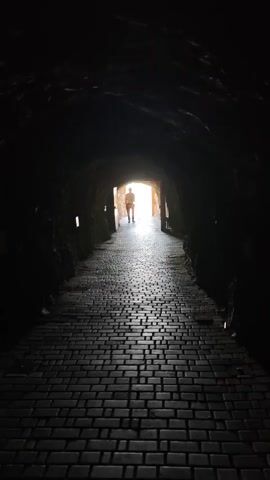 A person walks down a dark tunnel towards a bright exit. The tunnel is made of rough-hewn rock and lit by small lights embedded in the walls. The floor is paved with dark bricks, creating a textured pathway. The person is silhouetted against the light, making their features indistinguishable. The overall scene evokes a sense of mystery and transition.