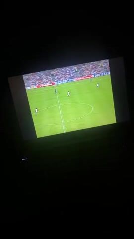 A football match is being displayed on a screen in a dark room. The screen shows a vibrant green pitch with several players in action. One player, identified by the commentator as Ronaldinho and wearing a blue and red striped jersey, is prominently featured dribbling the ball. Other players are visible in white and blue jerseys. In the background, stadium stands with spectators are partially visible. Advertising hoardings around the pitch display text such as "VODAFONE" and "XACOBEO." Below the screen, the top portion of what appears to be a laptop or keyboard is dimly visible. A commentator's voice, speaking French, can be heard in the background. The room is dark, suggesting it is nighttime or has been dimmed to enhance the viewing experience.