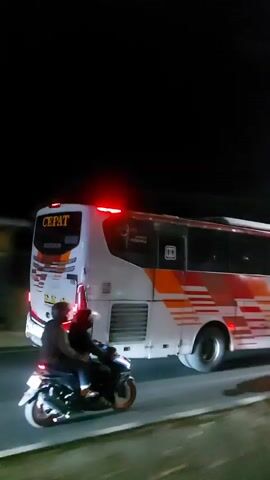 A white bus with orange and red racing stripes travels down a road at night. The bus has red lights flashing on its roof. A motorcycle with two people on it is traveling alongside the bus. The road is dark and appears to be a highway.