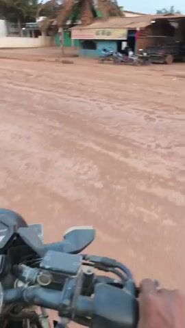 The video begins with a first-person perspective from someone riding a motorcycle. The rider's hands are visible on the handlebars. The scene is a dirt road with some shops or buildings lining the sides. Several motorcycles and a small truck are parked in front of the buildings. A palm tree is prominent in the background, along with some other trees and vegetation. The sky is overcast. The rider appears to be moving forward, and the camera pans slightly, indicating movement. The buildings have various signs, one of which appears to be for "Warung Mamak Ratna". There is also a tire and some debris on the ground. The overall impression is a rural or semi-urban setting with modest structures. The lighting suggests it might be early morning or late afternoon.