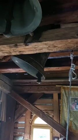 The video captures the interior of what appears to be a wooden bell tower or belfry. Two large bells are visible, suspended from thick wooden beams. The bell in the foreground is larger and hangs lower, its mouth open downwards. The second bell, slightly smaller and partially obscured, is positioned above and to the left. The wooden structure of the tower is rough-hewn and aged, with exposed beams and cross-supports. A rope hangs down near the right side of the frame, likely for operating the bells. To the right, a decorative golden cloth with an image of a religious figure is hanging on the wall. The overall lighting is dim, suggesting an indoor, possibly shaded environment. The sounds captured are the resonant ringing of the bells.
