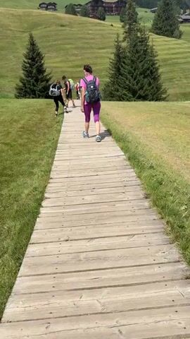The video shows a wooden boardwalk path leading up a grassy hill. There are people walking on the path. In the background, there are trees and buildings on the side of the hill, with a ski lift visible on the very top of the hill. The sky is cloudy.
