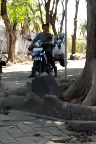 A person is seated on a motorcycle, holding a cigarette in their right hand and appearing to smoke it. The motorcycle is blue and white, with plastic bags secured to the rear. The person is wearing dark clothing. They are positioned on a paved area with cobblestone-like stones and large tree roots visible in the foreground. In the background, there are more trees and a wall. The lighting suggests it is daytime.