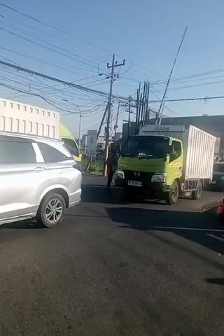 This video captures a bustling street scene in Gedangan, Indonesia, under a clear blue sky, suggesting it is daytime. The foreground is dominated by a silver SUV, its rear visible as it sits on the asphalt road. To the right, a green truck with a white cargo container is turning. Further back, another green truck is visible, and behind that, a white container truck is stationary. A dark-colored car is also present, and a person on a motorcycle is partially visible, suggesting active traffic. Power lines crisscross the sky, indicating an urban or semi-urban environment. A sign with "SOA019" is visible on a building in the background. The overall impression is one of everyday activity and movement in a populated area, likely a commercial or industrial zone given the presence of large trucks. The sunlight casts distinct shadows on the road, further confirming it's a bright day.