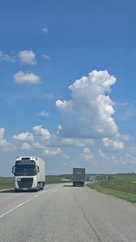 A video taken from a moving vehicle on a sunny day shows a highway in Panovka, Russia. The sky is a vibrant blue with fluffy white clouds, one of which is strikingly shaped like a rabbit. The road stretches into the distance, with green fields flanking either side. Several trucks are visible on the road; one large white trailer truck is in the foreground on the left, and a dark gray trailer truck with the "DSV" logo clearly visible is ahead in the right lane. A few other vehicles can be seen further down the road. The overall scene suggests a clear and pleasant day for travel.