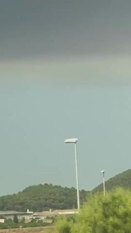 The video shows a landscape with rolling green hills covered in trees under a hazy, overcast sky. In the foreground, a few scattered buildings or structures are visible near the base of the hills. A tall, slender lamppost stands to the right of the frame. The camera appears to be moving slightly, possibly from a vehicle, as indicated by the blur of some vegetation in the lower left portion of the frame in the last few seconds. The overall atmosphere is muted and somewhat somber due to the cloudy conditions.