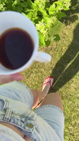 A person's hand holds a white mug filled with dark liquid, likely coffee, against a backdrop of green foliage and grass. The person is wearing denim shorts and pink flip-flops, suggesting an outdoor setting on a sunny day. The camera angle is a first-person perspective, focusing on the mug and the immediate surroundings. The video appears to capture a moment of relaxation or enjoyment in nature.