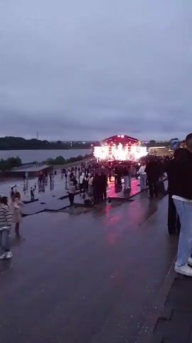 A wide shot captures an outdoor event with a stage set up in the background, illuminated by bright lights. A body of water is visible to the left of the frame. A crowd of people stands on a wet, paved area, some holding umbrellas, suggesting it might be raining or has recently rained. The sky is overcast. People are milling around, some looking towards the stage, while others are engaged in conversation or walking. The atmosphere appears to be that of a concert or festival.