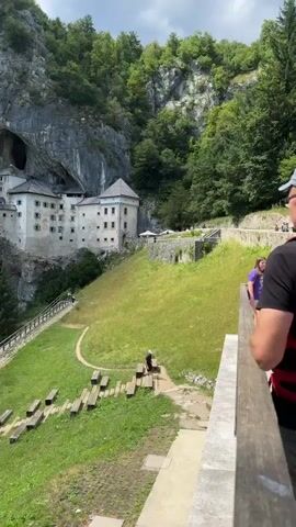 The video shows a medieval castle built into the side of a large cliff face. The castle is made of white stone with multiple levels and windows. A large cave opening is visible above and behind the castle. In the foreground, there is a grassy slope with a wooden walkway and seating area. Several people are visible walking around the grounds. The sky is partly cloudy and the overall environment appears to be a natural, somewhat mountainous landscape with dense green trees covering the upper parts of the cliff.