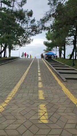 A paved walkway with yellow markings slopes upwards, lined by trees. On the left, a set of stairs ascends parallel to the walkway. Several people are walking up the walkway, some carrying bags. On the right, another set of stairs leads up, and there is a blue umbrella and a small stand or kiosk. The sky is cloudy.