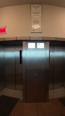 The video shows two elevators, each with a digital display above the door. The display on the left shows -3.7 and the display on the right shows increasing numbers starting at 140.0 and eventually reaching 155.0. The elevators are silver and metallic, and the floor is covered in brown and orange speckled tiles. A poster with diagrams and text in German hangs above the elevators. A voice can be heard speaking in the background.