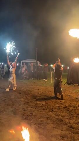 The video shows a nighttime performance with fire dancers. In the foreground, two people are performing with fire. One person on the left is holding a sparkler that is emitting a spray of bright white sparks upwards. This person is wearing a white dress and white boots. The other person on the right is holding a flaming object that is creating a large plume of fire. This person is wearing a darker outfit with what appears to be a headdress. There are several other fires burning on the ground, and in the background, there are blurred figures of spectators and tents or structures, indicating an outdoor event. The ground appears to be sandy or dirt. The overall atmosphere is dark with bright light sources from the fire and sparks, and some smoky haze.
