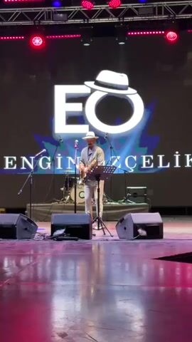 The video shows a man playing an electric guitar on a stage. He is wearing a light-colored hat, a light-colored shirt, and light-colored pants. He is standing in front of a large screen with the letters "EO" and a hat logo. There are microphones and speakers on the stage, and a drum set is visible in the background. The lighting is dim, with spotlights on the performer. The man appears to be in the middle of a performance, holding the guitar and looking towards the audience. The video is short and focuses on the performer.