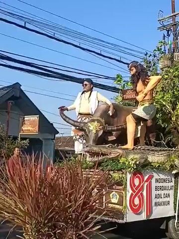 The video shows a parade float moving down a street. The float is decorated with large statues of a man riding a buffalo, which is standing on top of a crocodile. The float also has a large "81" graphic and text that says "INDONESIA BERDAULAT ADIL DAN MAKMUR". There are people on the float and walking alongside it, some of whom appear to be wearing traditional clothing. The background shows buildings, trees, power lines, and a blue sky. The float is on a truck and appears to be part of a celebration.