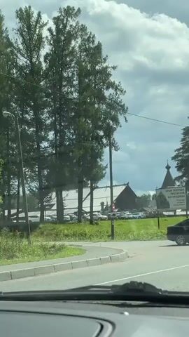 The video shows a road and trees with a large wooden building in the background. A sign in front of the building reads "ХРАМ ПОКРОВА ПРЕСВЯТОЙ БОГОРОДИЦЫ" which translates to "Church of the Intercession of the Most Holy Mother of God". There are cars parked in front of the building and a black car driving on the road. The sky is cloudy.