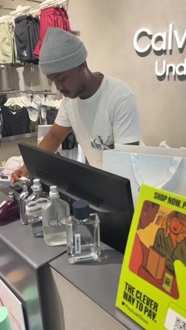In an indoor shot, a man wearing a gray beanie and a white Calvin Klein t-shirt stands behind a counter in a Calvin Klein store. He is looking at a computer monitor and appears to be working. On the counter, there are several bottles of Calvin Klein cologne. To the right of the monitor is a sign that says "SHOP NOW. PAY...", advertising a payment service. Behind him, there is a clothing rack with various items of clothing, including shorts and shirts.