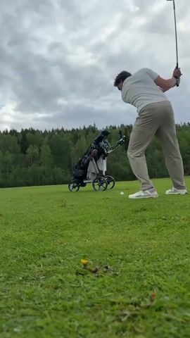 A man is swinging a golf club on a grassy golf course. He is wearing a white polo shirt, tan pants, white golf shoes, and a golf glove on his left hand. His golf bag is in a push cart behind him. The sky is cloudy and there are trees in the background. The golfer is facing away from the camera as he swings.