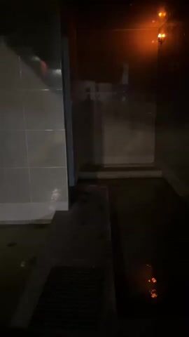 The video shows an interior space with tiled walls and a floor that appears to be wet or submerged. The lighting is dim at the beginning, with some orange glows visible, and then transitions to brighter, more even lighting. In the center of the room, there is a raised concrete ledge or platform, and further into the space, a water faucet is visible on the wall. The floor has a greenish tint, suggesting the presence of water and possibly algae or mildew. There is a drain grate visible on the floor. The overall impression is that of a utility area or a space designed to be washed down, possibly a shower or a service area. No people or specific actions are depicted beyond the camera's movement.